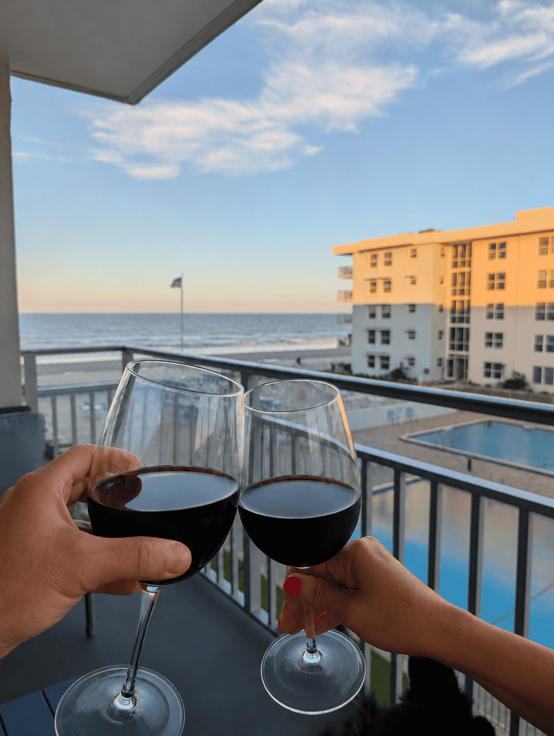 Two people toast with glasses of red wine on a balcony overlooking the ocean and pool at sunset.
